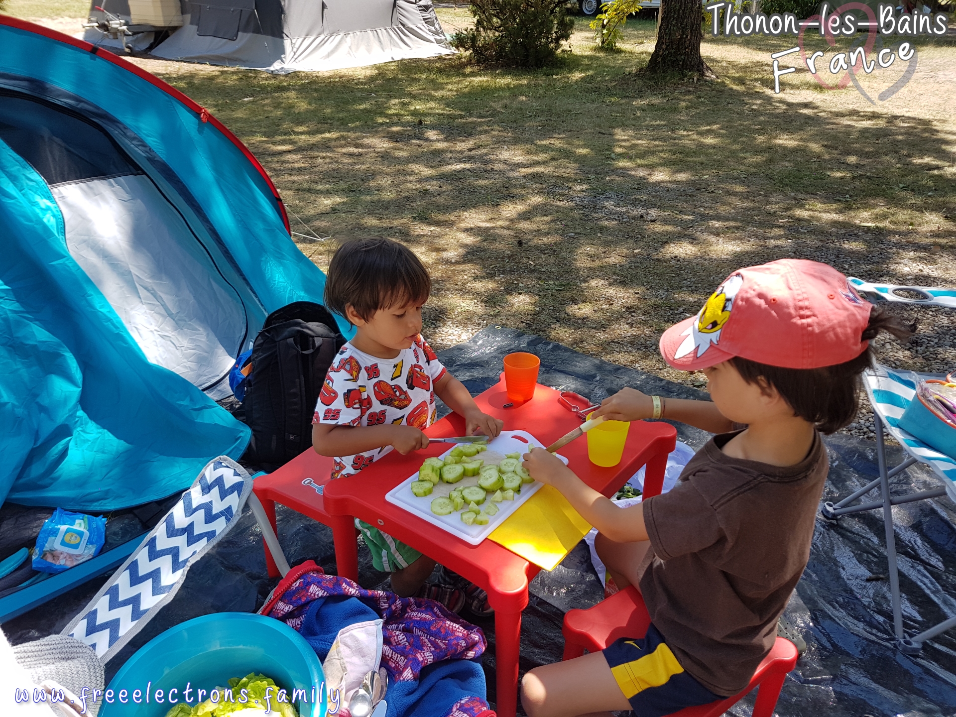 #FreeElectrons.Family - camping road trip Europe, Lake Geneva.

Two young kids at work, cutting cucumbers on a red toy table in front of a tent in a shaded camping site.

Text reads: Thonon-les-Bains France (with 2 opaque interlocking hearts behind the text) and www.freeelectrons.com.