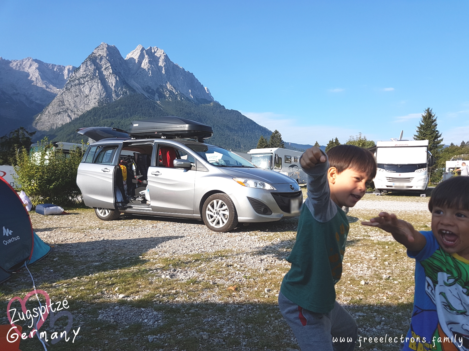 #FreeElectrons.Family - camping road trip Europe, Quechua and Mazda, Germany.

Kids having fun at a camping site with rocky mountain peaks in the background.  Behind them are RVs, a silver Mazda MPV 5 with a black Thule Roofbox, and part of a Quechua tent.

Postcard-like text reads: Zugspitze, Germany with an opaque image of two hearts behind the text and www.freeelectrons.com.

